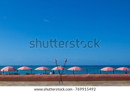 Pink umbrellas at Waikiki beach at the pacific ocean with blue skies in the background and a paved patio. Two tiki torches