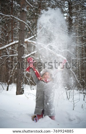 blond woman on vacation throwing snow in air in forest with a nice smile. Cute girl wear red scarf and knitted mittens , gray fur hat and stylish coat. Good weather and snowfall. Full length female