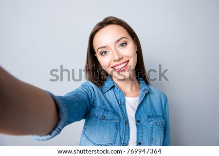 Selfie mania. Close up of confident brunette girl  with beaming smile in denim oufit taking a self-portrait  on her mobilephone and  standing over grey background