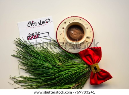 A cup of coffee and Christmas loading text on a note on white wooden table and green tree branch with a red bow