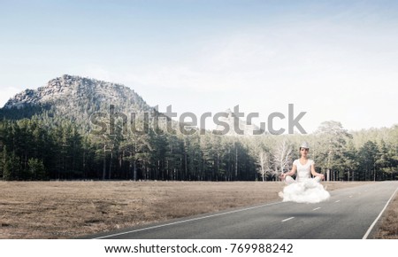 Young woman keeping eyes closed and looking concentrated while meditating on cloud above the road with beautiful and breathtaking landscape on background.