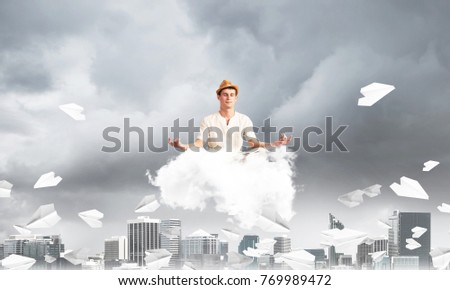 Man in white clothing keeping eyes closed and looking concentrated while meditating on cloud among flying paper planes with cityscape view on background.