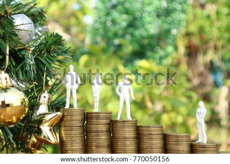 Miniature businessman and woman stand on level rolls ladder of gold coin money lead to Christmas tree decorated with gold stars and silver balls in blur natural tree bright light