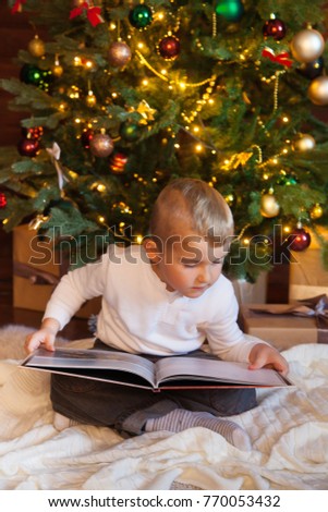 The boy looks at the book under the Christmas tree.