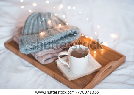 Cup of coffee on open book staying on wooden tray with knitted clothes close up over Christmas lights. Good morning. Winter season. 