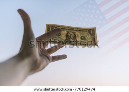 One dollar in hand with USA flag over sky background.