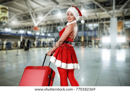 Slim young woman in red hat and santa claus dress with big red suitcase. Airport interior. Christmas travel time. 