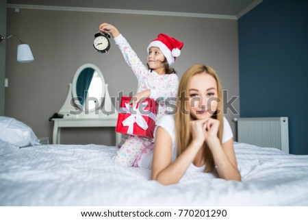 Mother lying in the morning bed with little daughter wearing in santa hat holding clock and santa gift and show that its time to celebrate Christmas. Christmas and New year