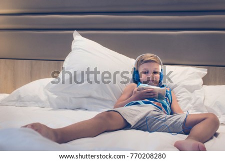 Little boy lying on bed and watching cartoons on smart phone. Smiling kid with headphones listening music while relaxing on the bed and using mobile phone. 