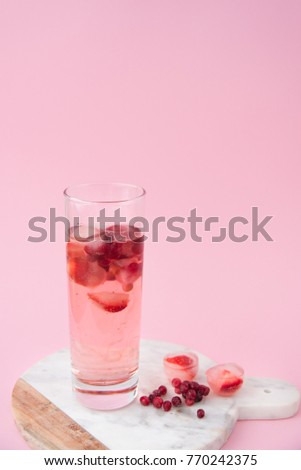 A glass of iced tea with frozen strawberry and lingonberry on a marble stand on pink monophonic background.