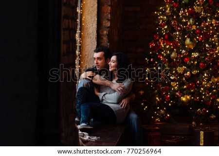 Couple of lovers hugs sitting on the windowsill in the Christmas loft studio. guy is hugging the girl. Garlands and New Year's lights. backdrop of a Christmas tree. Brick wall.