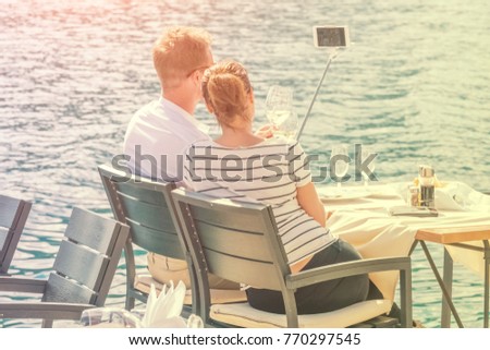 Young couple is sitting at a table by the sea with glasses of wine and is photographed on the phone