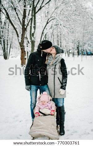 Mom kissing Dad and daughter stand outdoors on the background winter. Happy family walks in the park. full length. Close up. Portrait baby in children's sled. Infant.