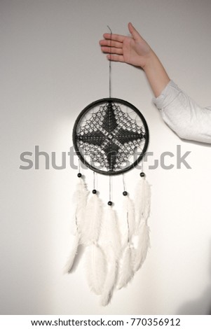 Black dream catcher with a round base, cobweb, crocheted, with a diamond, large white goose feathers and black beads is holding a girl against a white wall background