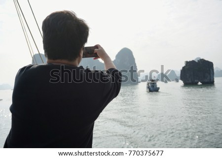 A man with short black hair wearing a long- sleeved black shirt using a smartphone to take photos on a sunny day of sailing boats, different shapes and sizes of rocks in Ha Long Bay in Vietnam.