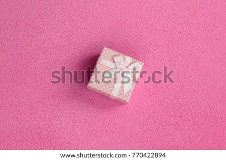 A small gift box in pink with a small bow lies on a blanket of soft and furry light pink fleece fabric. Packing for a gift to your lovely girlfriend