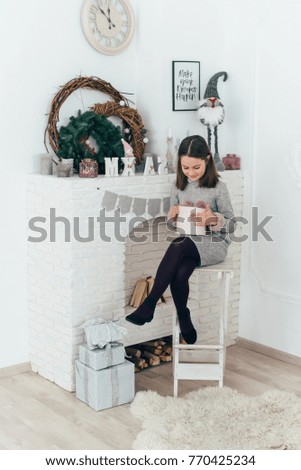 A girl sits at the fireplace and holds a box with a gift in her hands