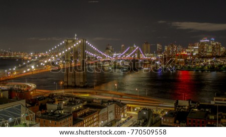 A view of the Brooklyn Bridge with the brightly lit neighborhood of Brooklyn Heights in the background. The sky is brightly lit with a noticable cloud and the river reflection is brightly lit.