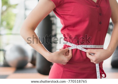 Young slim woman measuring her waist by measure tape after a diet with accessory in sporty gym as background. Royalty-Free Stock Photo #770518765