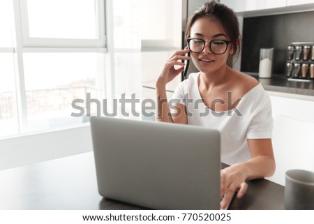 Photo of happy young woman sitting at the kitchen in home. Looking aside using laptop talking by mobile phone.