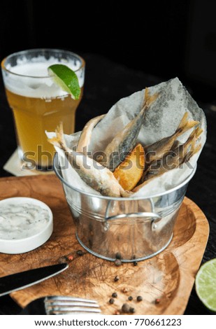 snack to beer - fried small fish with potatoes and sauce on a wooden plate with a glass of light beer