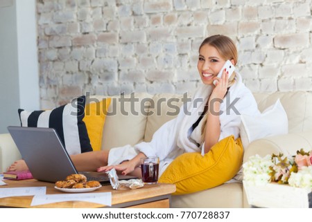 Female freelancer in a white coat clothing working remotly from her dining table in the morning. Houses on a sofa on a white brick wall background. Architect, blueprints. talking on the phone.