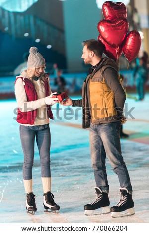 side view of young man with balloons presenting gift box to girlfriend at st valentines day on rink  