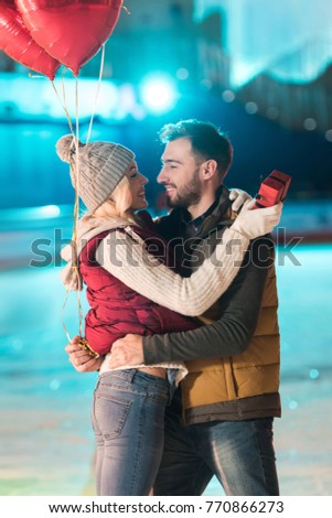 happy young couple with gift box and heart shaped balloons looking at each other on skating rink 