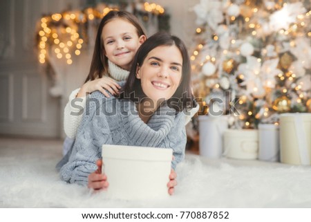 Indoor shot of mother and daughter have fun together, share presents, being in room decorated with garlands and Christmas tree, have joyful expressions, enjoy weekends and winter holidays