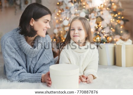 Winter and celebration concept. Pretty lovely blue eyed small female child in knitted white sweater and brunette female have good rest at home, spend time near adorned Christmas tree