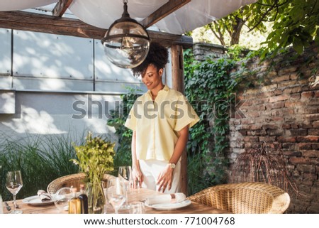 Beautiful African woman arranging dishware on terrace table for hosting the celebration.