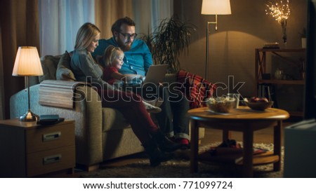 Long Shot of a Father, Mother and Little Girl Watching TV. They Sit on a Sofa in Their Cozy Living Room, Father Holds Laptop on His Knees. It's Evening.