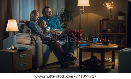 Long Shot of a Father, Mother and Little Girl Watching TV. They Sit on a Sofa in Their Cozy Living Room and Eat Popcorn. It's Evening. Royalty-Free Stock Photo #771099727