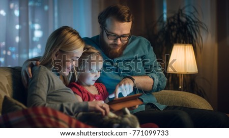 Father, Mother and Little Daughter Reading Children's Book on a Sofa in the Living Room. It's Evening.