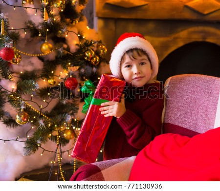 Beautiful little brunet boy, has happy serious face, brown eyes, has red Christmas hat Santa Claus, hold gift box. Portrait family holiday. Close up. Winter background. Tree toys. Night light.