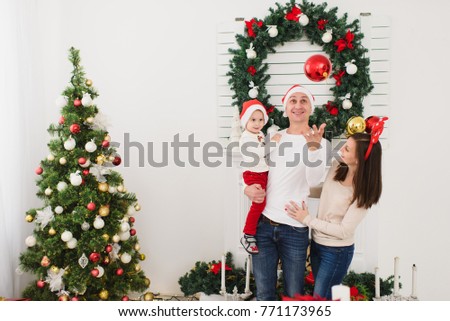 Happy parents with cute little son in hat. Child boy with big red tree toy ball standing in decorated New Year light room at home on the background of Christmas wreath. Family, holiday 2018 concept