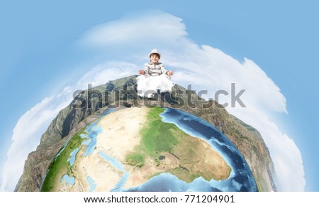 Young little boy keeping eyes closed and looking concentrated while meditating on clouds in the air with panoramic view of Earth globe on background. Elements of this image are furnished by NASA