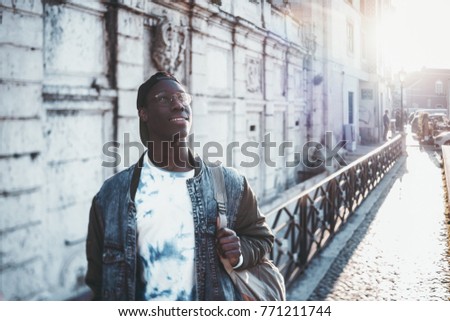 Handsome African student boy in a jean jacket and round glasses is looking upside while standing outdoor; male black undergraduate young guy on his way to university with copy space place for text Royalty-Free Stock Photo #771211744
