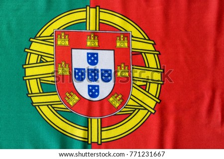 A Portuguese flag is pictured as it lays on a flat surface. The Coat of Arms in between the bold red and green colors is a great symbol for the country of Portugal.