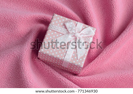 A small gift box in pink with a small bow lies on a blanket of soft and furry light pink fleece fabric with a lot of relief folds. Packing for a gift to your lovely girlfriend