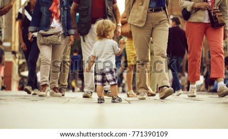 Photo back view of cute fair-haired blond kid tiny little child baby boy eating bun standing on flag-stone pavement in crowd cityscape on blurred grey background, horizontal picture
