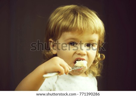Closeup portrait view of one adorable cute small baby boy with blonde hair eating healthy food of porridge or coocked semolina with spoon in hand indoor, horizontal picture