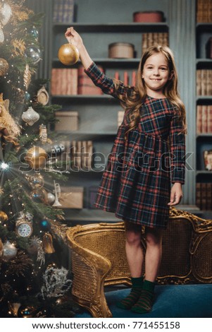 Pretty girl with charming smile stands on armchair, decorates New Year tree alone, has good mood, feels proud to do it by herself. Small kid prepares for Christmas. Childhood and holidays concept