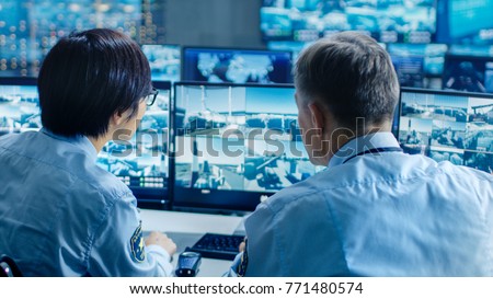 In the Security Control Room Two Officers Monitoring Multiple Screens for Suspicious Activities, They Report any Unauthorised Activities with Walkie-Talkie. They  Guard Object of National Importance. Royalty-Free Stock Photo #771480574