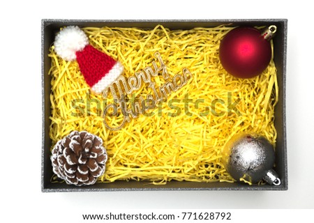 Merry christmas text and fur hat, ball, pine cone in black gift box with yellow packaging material
