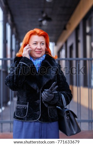 A woman in a fur coat and talking on the phone is standing in the street on a blurred background