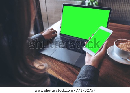 Mockup image of business woman holding mobile phone with blank green screen while using laptop on wooden table in cafe