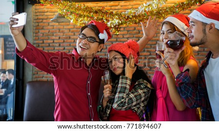 Group of cheerful Asian freind, handsome man and beautiful woman wearig Santa hat, holding smart phone and making selfie at Christmas party in restuarant.