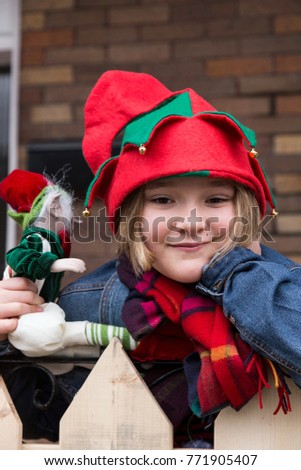 Vertical closeup of beautiful round faced pink cheeked little girl with mischievous expression wearing elf hat and holding an elf toy
