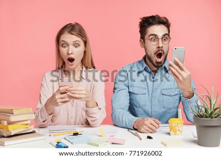 Portrait of shocked terrified male and female stare at mobile phones, type messages or read shocking news, sit next to each other at work place, isolated over pink background. Suprised students indoor Royalty-Free Stock Photo #771926002
